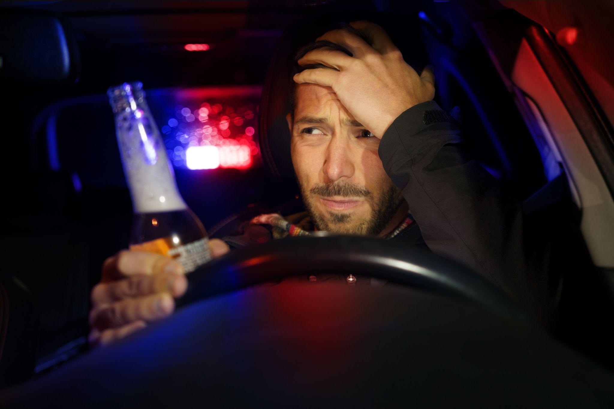 Man holding an open beer bottle while sitting behind the wheel of a car. He looks exasperated as red and blue lights flash behind him.